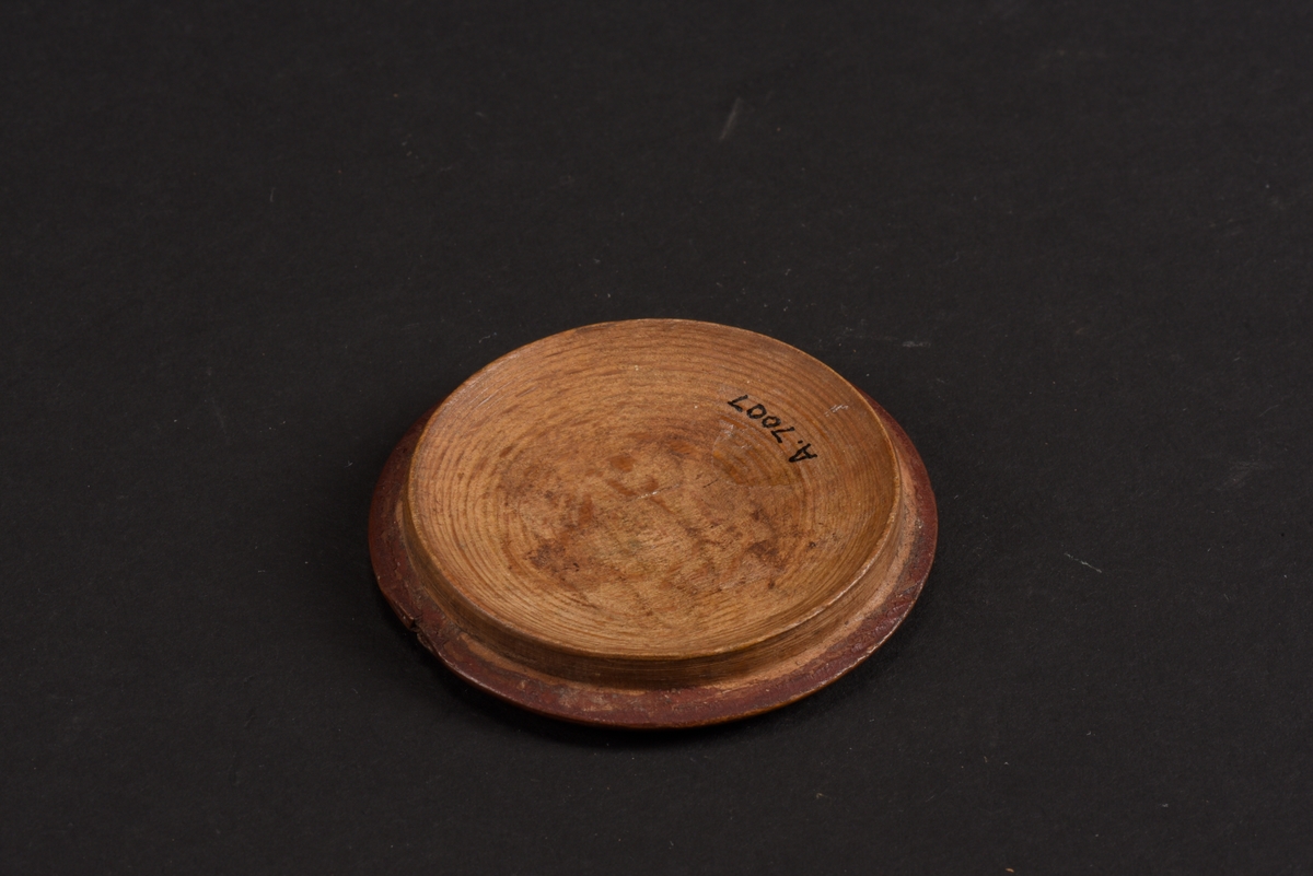 Rund, svarvad, ask med lock tillverkad av bok. Asken är målad på utsidan i rött med gula mönster; runt asken ett sicksackmönster och på locket ett årtal, 1760, samt en stiliserad lilja. Asken är skadad och en bit saknas.
