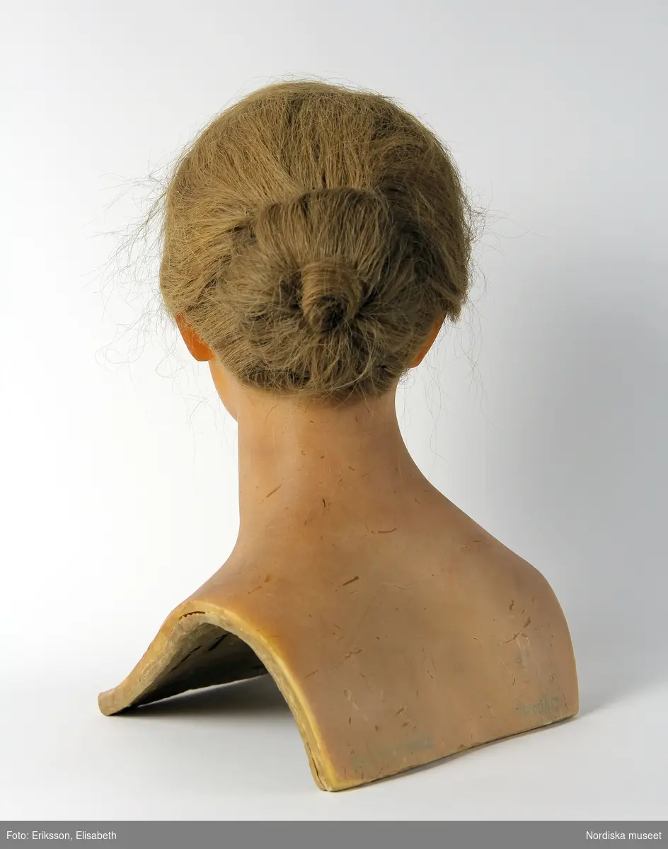 Skyltdockshuvud, kvinna, av vax med riktigt hår, ögonfransar och ögon av glas. Med hals och övre del av torso. På baksidan av torso text "Paul Baschwitz / Berlin C". Blont hår friserat i knut (hårnålar ingår ej i förvärvet).
Anm. spricka vid höger och vänster öga och vax gulnat.
/Helen Persson 2018-02-13