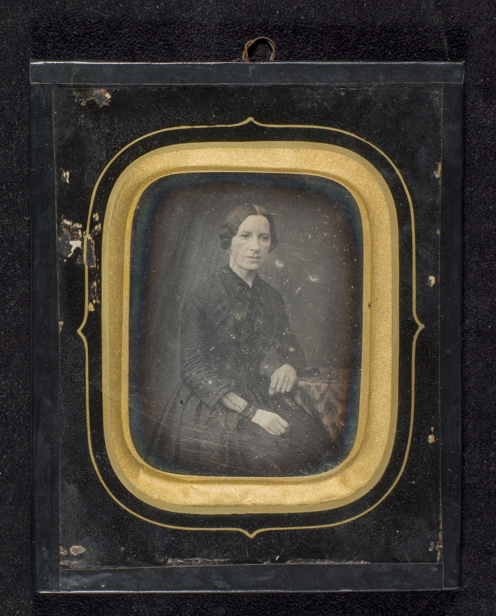 Daguerreotypi av sortkledd kvinne sittende ved et bord.