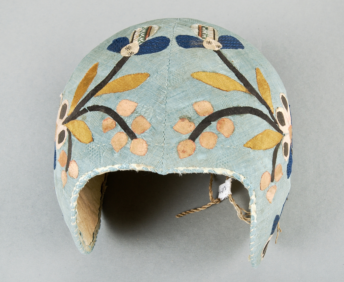 Bindmössa av blå/grön mönstrad siden, Påklistrade applikationer i form av blommor, stjälkar och blad i brunt, rosa, blått, beige och ett möstrat/randigt tyg. Fodrad med beige tuskaftat lintyg. Hoprynkad baktill i sidorna, försedd med dragband.