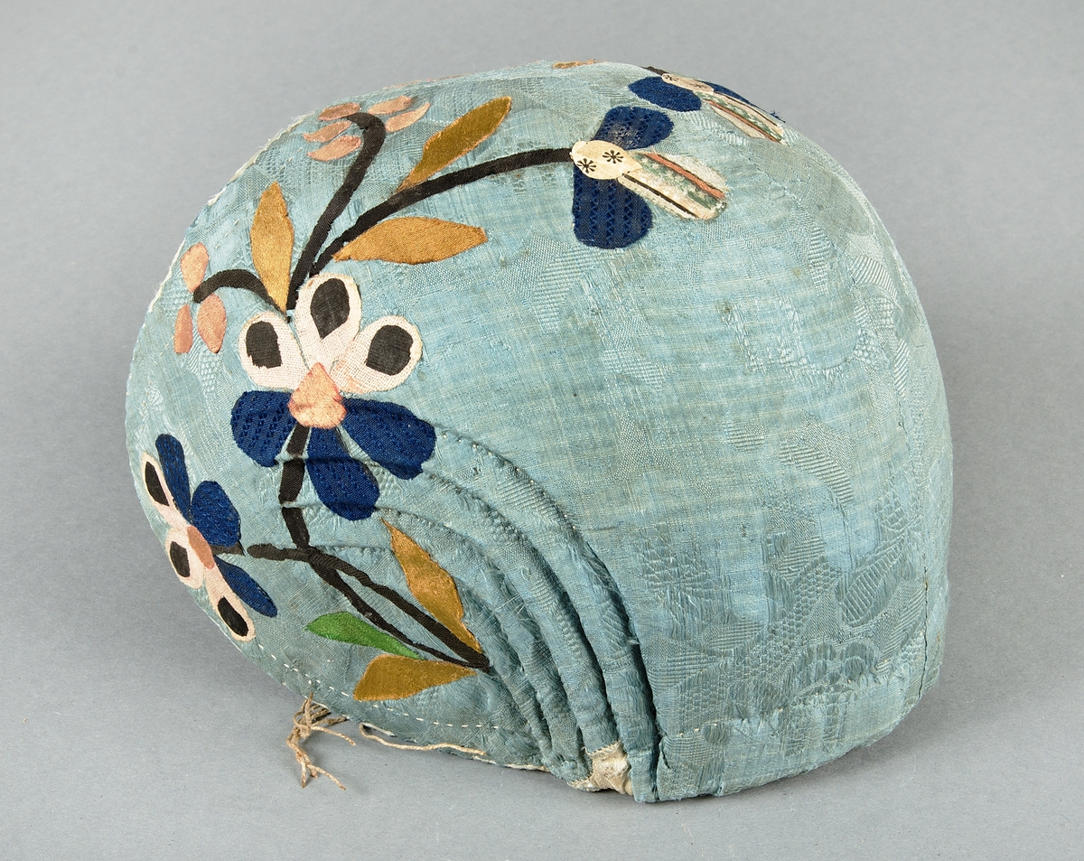 Bindmössa av blå/grön mönstrad siden, Påklistrade applikationer i form av blommor, stjälkar och blad i brunt, rosa, blått, beige och ett möstrat/randigt tyg. Fodrad med beige tuskaftat lintyg. Hoprynkad baktill i sidorna, försedd med dragband.