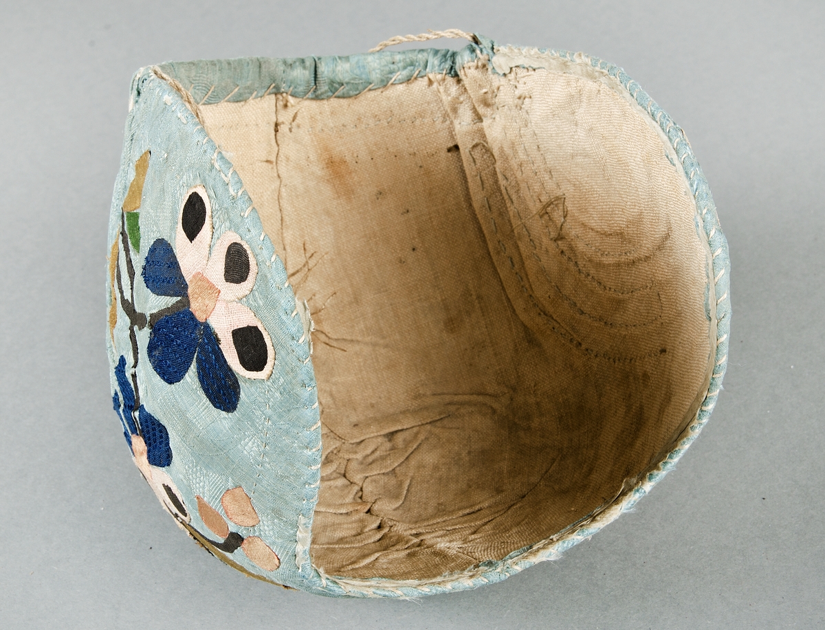 Bindmössa av blå/grön mönstrad siden, Påklistrade applikationer i form av blommor, stjälkar och blad i brunt, rosa, blått, beige och ett möstrat/randigt tyg. Fodrad med beige tuskaftat lintyg. Hoprynkad baktill i sidorna, försedd med dragband.