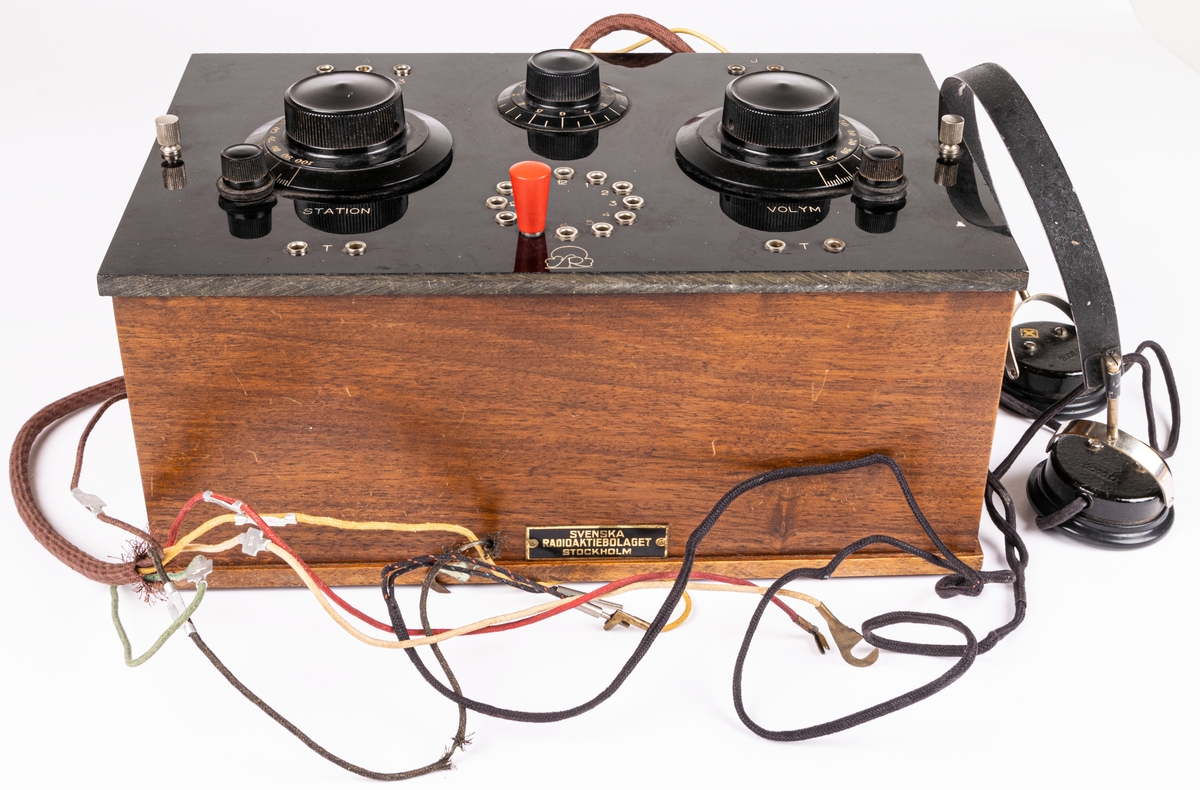 Batteridriven 2 rörs radioapparat. Lådan av svart och brunt trä. 2 par hörlurar. Diverse sladdar. På lådans sida en metallbricka "SVENSKA RADIOAKTIEBOLAGET STOCKHOLM" På undersidan provsedel med text: "...RADIOLA M60...".