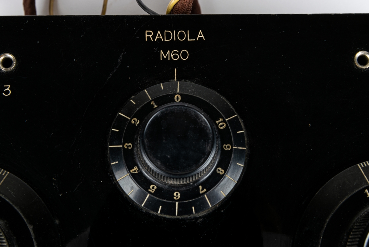 Batteridriven 2 rörs radioapparat. Lådan av svart och brunt trä. 2 par hörlurar. Diverse sladdar. På lådans sida en metallbricka "SVENSKA RADIOAKTIEBOLAGET STOCKHOLM" På undersidan provsedel med text: "...RADIOLA M60...".