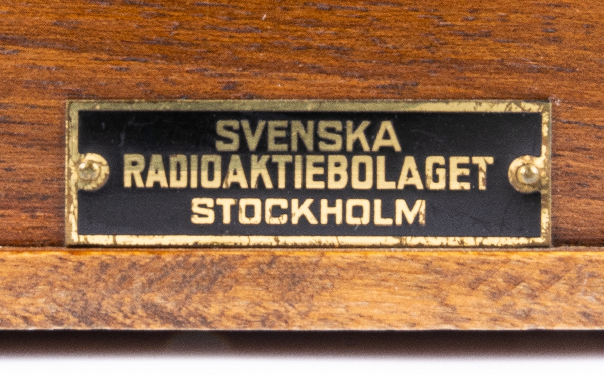 Batteridriven 2 rörs radioapparat. Lådan av svart och brunt trä. 2 par hörlurar. Diverse sladdar. På lådans sida en metallbricka "SVENSKA RADIOAKTIEBOLAGET STOCKHOLM" På undersidan provsedel med text: "...RADIOLA M60...".
