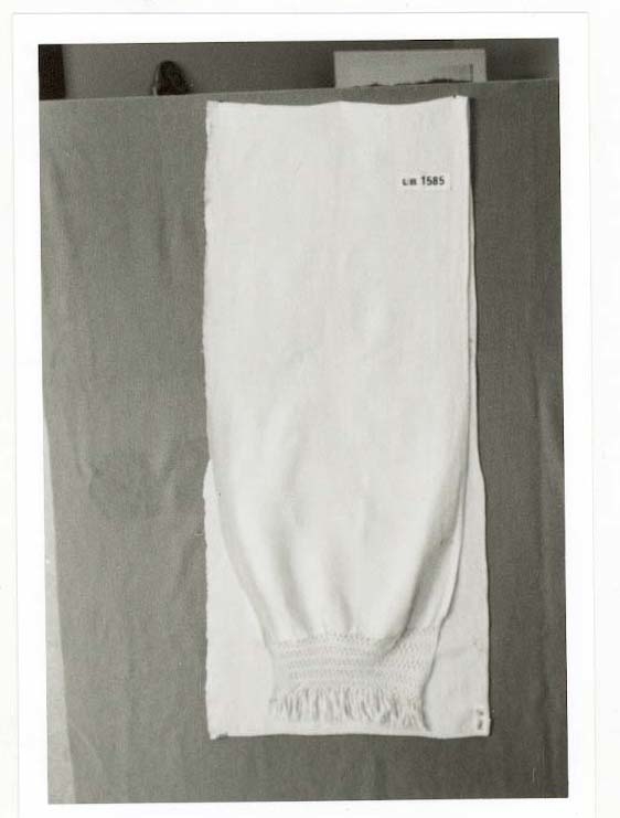 Grautspannklede i kvit, heimvove lin, ca 16 trådar per cm. blonde i firfletting på den eine kortsida. Små duskar i enden av blonda.