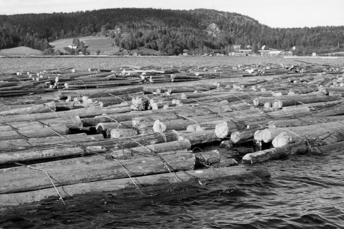 Tømmerslep på innsjøen Visterflo, like utenfor lenseanlegget ved Eidet i Tune i Østfold høsten 1953.  Fotografiet er tatt med to slep av barket tømmer på vannet i forgrunnen, og til høyre i bakgrunnen skimter vi lenseanlegget inne ved strand.  I forgrunnen ser vi hvordan slepene var sammensatt av "moser" (bunter) som ble holdt i hop av tre-fire "bind" - vaiere med en åttetallsformet jernring ("kaus") i den ene enden og en kjettingstump som kunne tres gjennom jernringen og "låses" i den andre. I sideretningen ble mosene holdt sammen av "tverrvirer".  Dessuten fantes det en kraftig vaier som gikk rundt slepet, langs ytterkantene, og som ble kalt for "slepeselen".  Lenseanlegget ved Eidet, der tømmertunnelen fra Glommas vestre løp kom ut av det bakenforliggende fjellet, skimtes til høyre i bakgrunnen. Fotografert 26. 09. 1953.