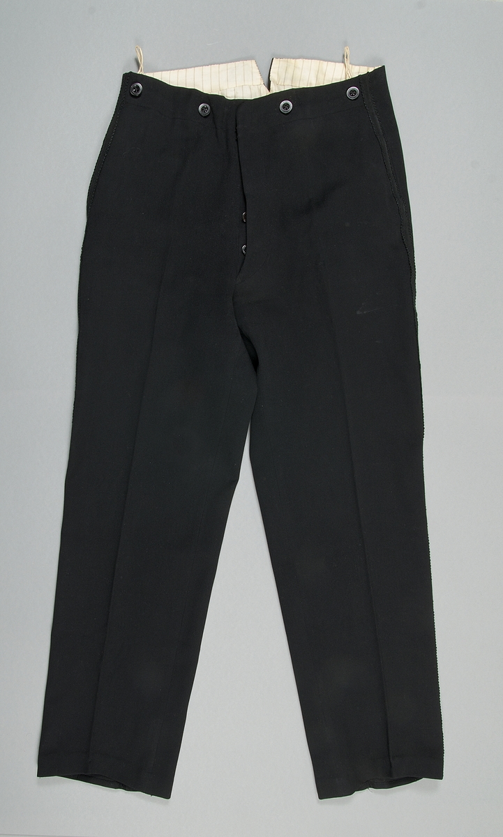 Byxa till smoking av svart kläde. Fodrad med randig, beige och svart, bomullssatin. Revärer i svart med mönster. Byxan stängs med knappa och hyska. Knapparna märkta "Georg Pettersson Rasbo", detsamma gäller knapparna för hängslen.