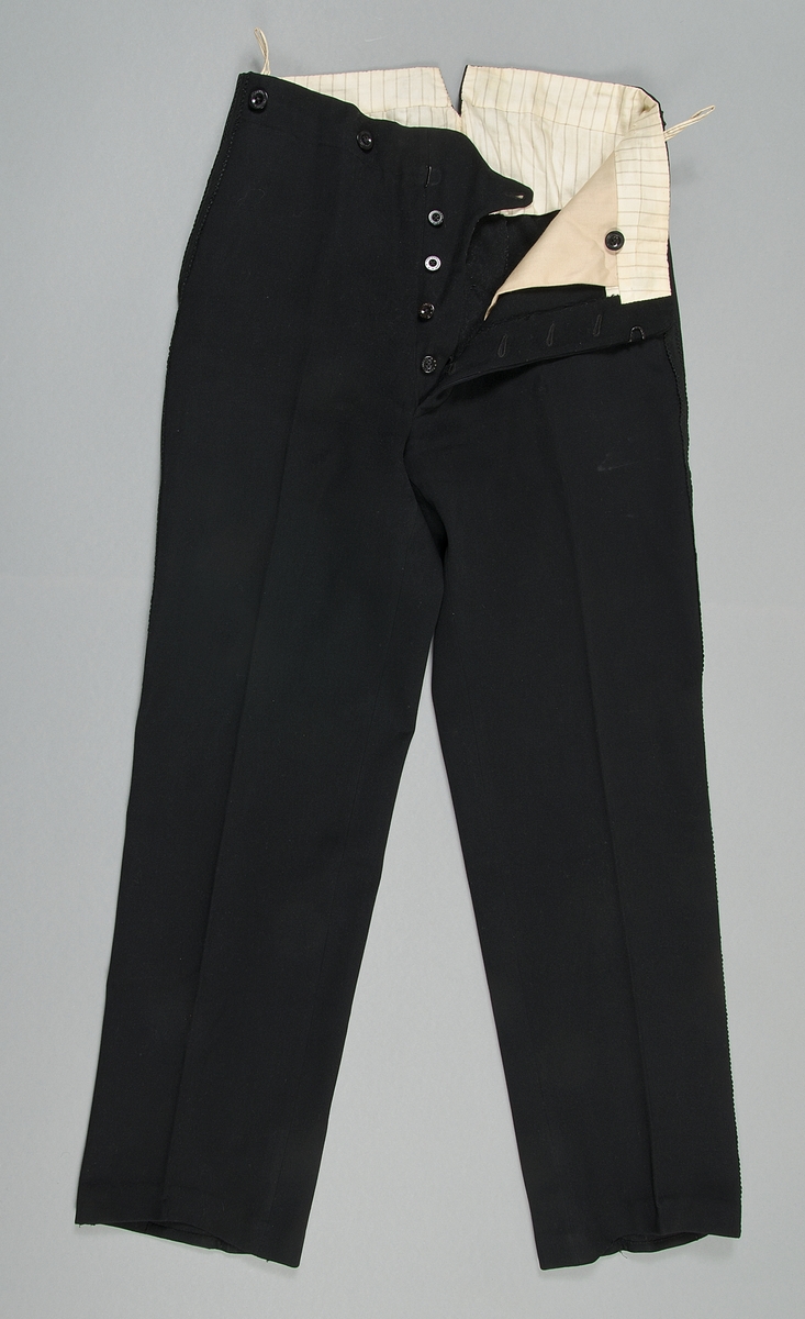 Byxa till smoking av svart kläde. Fodrad med randig, beige och svart, bomullssatin. Revärer i svart med mönster. Byxan stängs med knappa och hyska. Knapparna märkta "Georg Pettersson Rasbo", detsamma gäller knapparna för hängslen.