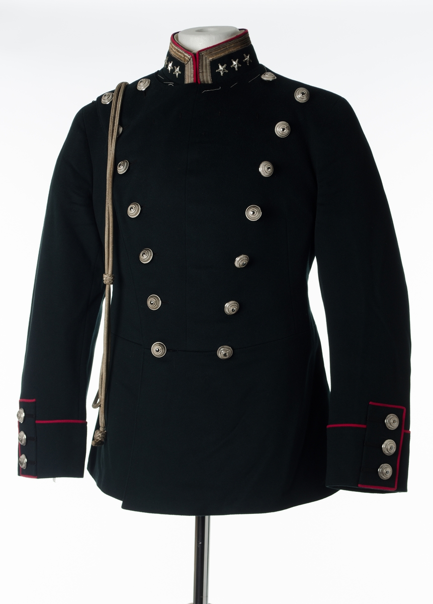 Dobbeltspent jakke til gallauniform i mørk grønn ull, med burgunderrøde detaljer og sølvknapper. Kraven er opprettstående og har ett sølvbånd og tre stjerner. Høyre skulder har to sydde hemper. Fronten har 16 knapper, festet med små plastkanpper på innsiden. Knappene er høye med fargene sølv og hvitt. Knappene har dekorative elementer som henspeiler på et hjul med et horn i midten. Seks små, sydde hemper øverst på venstre side. Baksiden har burgunderrød kantebånd i ryggen, splitt og tre par knapper ved splitten. Splitten er sydd sammen. Innerlomme på venstre side. Nederst på ermene er det tre bånd med knapp og burgunderrøde kantbånd. Innvendig er jakken helforet med sort bomullsstoff og lyst stripete bomullsstoff i ermene. Opphengshemper i ermene. Fronten er vattert. Innvendig belte med tre flate hekter i metall i midjen. Løssnipp er festet på metallnagler. Stivet løssnipp i hvit bomull med fem hull for feste til metallnagler på innsiden av opprettstående krave. Hvite tråklesting i kraven.
Dobbel sølvfarget snor henger på skrå over jakken.