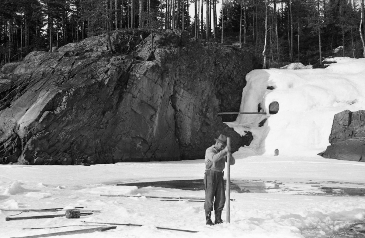 Forberedelse til isskytning eller issprenging i Forssvika nedenfor Braskerudfossen eller Braskereidfossen i Glomma i Våler i Hedmark. Her ser vi en mann som stakk en stolpe ned i et hull i isen, der det antakelig skulle plasseres en ladning. Flere stolper lå på isen bak mannen. Målet med dette arbeidet var antakelig å etablere en isfri kanal, der fløtingstømmeret kunne passere.