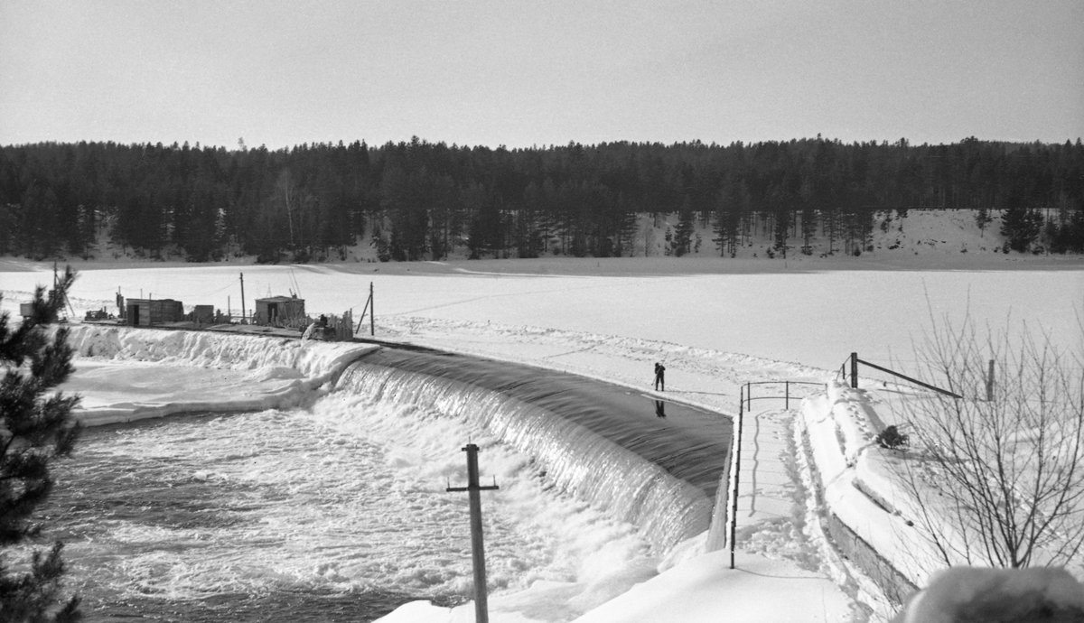 Anleggsarbeid på den midtre delen av den drøyt 300 meter lange terskeldammen ved Skjefstadfossen i Heradsbygda i Elverum, Hedmark. Fotografiet ble tatt i slutten av februar måned i 1942. Da var elva, med unntak av det østre løpet nedenfor dammen (i forgrunnen, der vannføringa var størst) is- og snødekt. Anleggsarbeiderne hadde rigget seg til med brakker ved den midtre delen av dammen (til venstre på dette bildet), og de hadde fått strømforsyning og oppkjørt veg ut til arbeidsstedet. Sannsynligvis skulle de forsterke dammen, som på dette tidspunktet var drøyt 30 år gammel. Slikt arbeid ble gjerne gjort i vintesesongen, for da var vannføringa i elva liten.