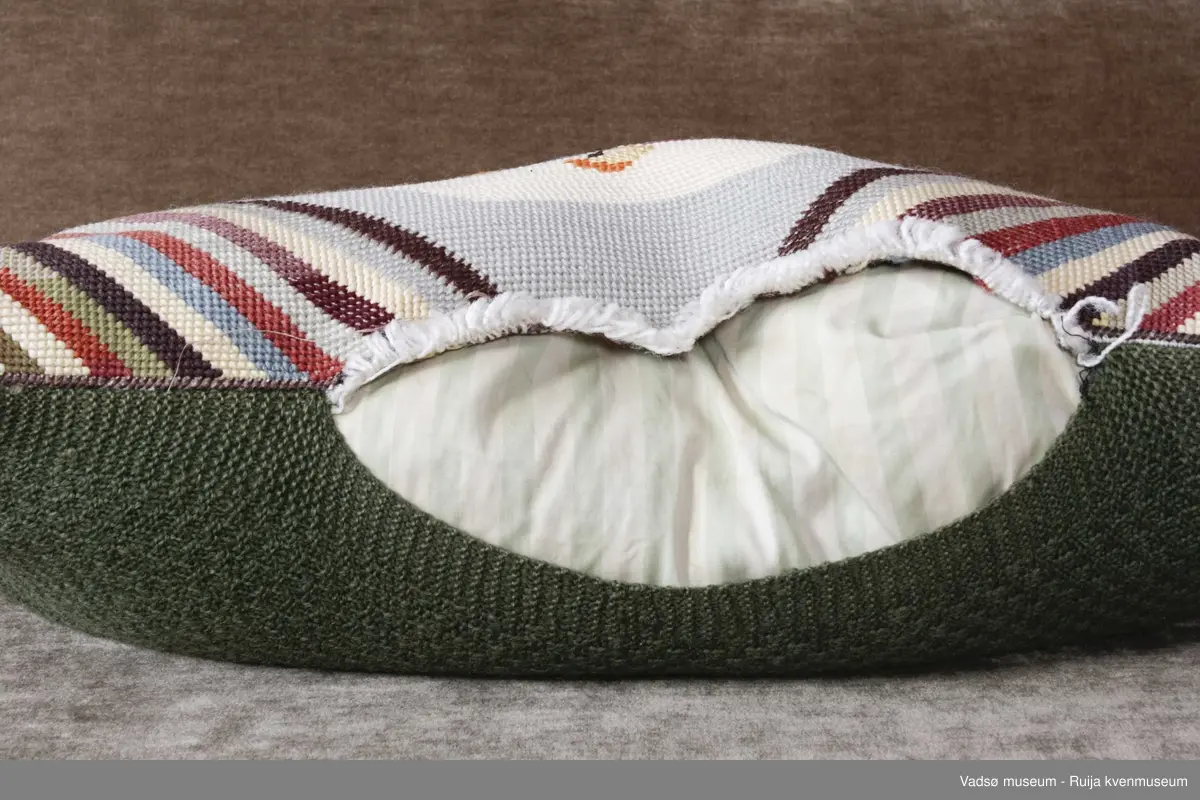 1 stk pute. Tidl. reg. brodert korssting på tysk sekk.Fra ca. 1942-43
Blomstermotiv i midten, striper diagonalt i hjørnene. Strikket grønn bakside.