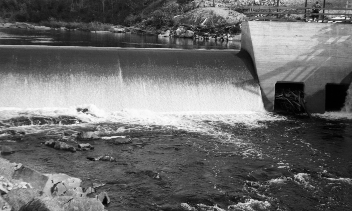 Kraftverksdammen ved Kvernfallet i elva Søndre Osa i Åmot, Hedmark, fotografert i 1941. Dette var en terskeldam, som skulle stuve opp vann til et turbinrør som gikk langs den søndre elvebredden til en kraftstasjon ved Brufallet, noen hundre meter lengre nede i vassdraget Dette fotoghrafiet er tatt i motstrøms retning, mot dammen, der det på dette tidspunktet, antakelig litt ut på sommeren, gikk en vannstrøm over den avrundete damkrona, som falt kvitskummende ned i det nedenforliggende elveløpet. Inntaket til turbinrøret befant seg i bassenget bak betongmuren til høyre i bildet. Til høyre i bakgrunnen skimter vi en mann ved et gjerde, som antakelig skulle skjerme det nevnte bassenget.