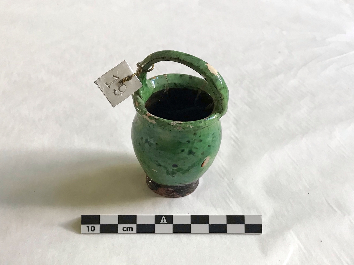 Liten grønn leirkrukke med handtak. Glasert. 

Har flere revner, delvist ødelagt. Har vært limet sammen.