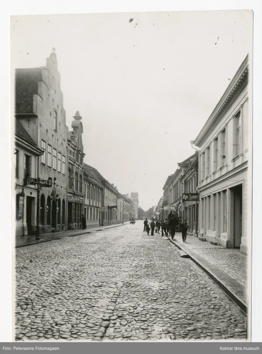 KLM 45133:1. Fotografi. Svartvitt. Motiv: Storgatan i Kalmar med kullerstenslagd gata, daterad till 1901. Folkliv på gatan med en man som klättrar på en stege. Stämplad: Peterssons Fotomagasin, Kalmar.