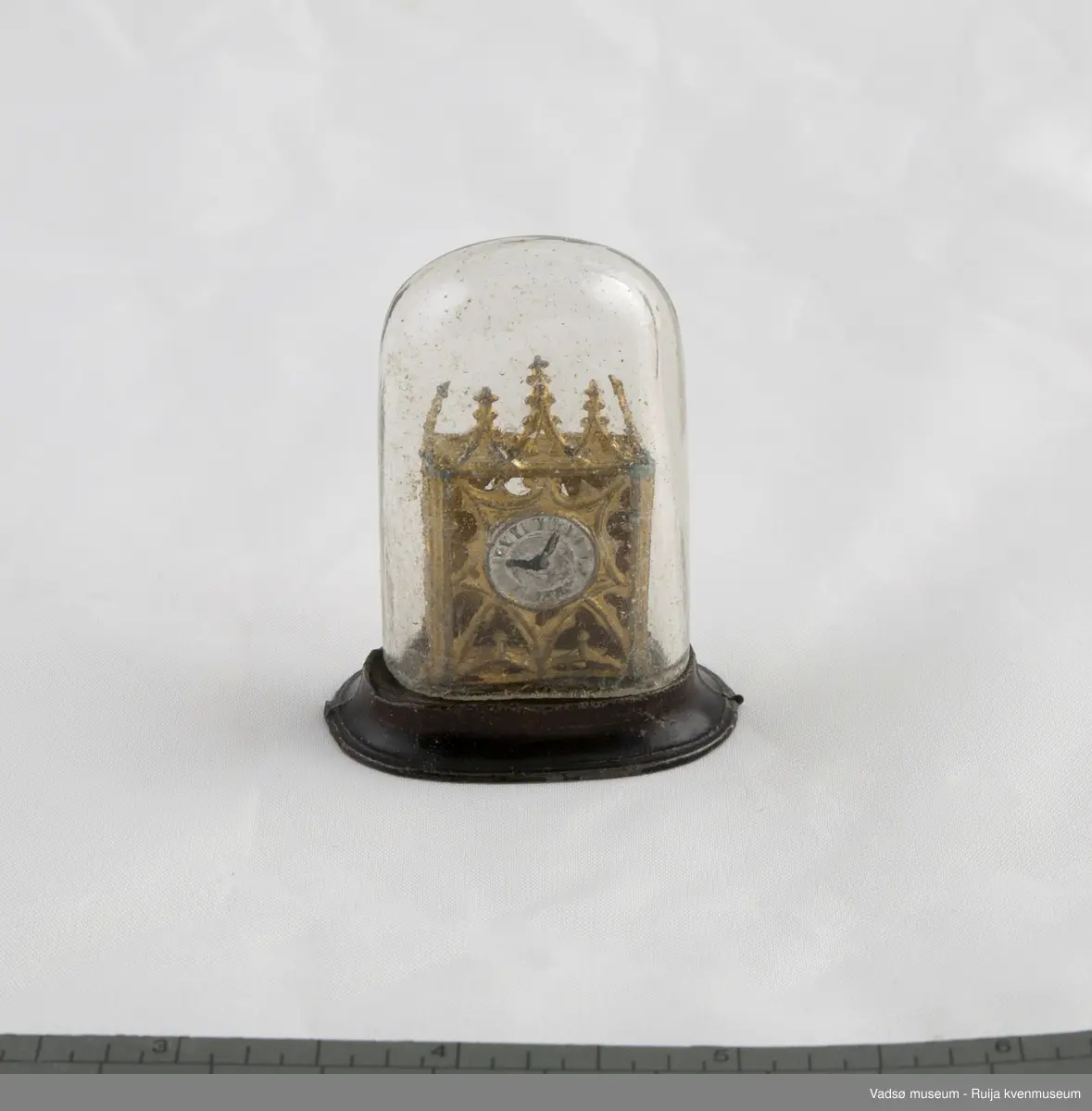 Dukkehusinventar. Forestiller bordklokke i metall. Rektangulært klokkehus med 5 spir i front. Klokkehus er loddet fast i oval metallsokkel som er noe bredere enn klokkehuset. Urskive i sølv og visere i svart. Klokka har en pynte/beskyttelseskuppel i tykt glass som kan tas av og på.