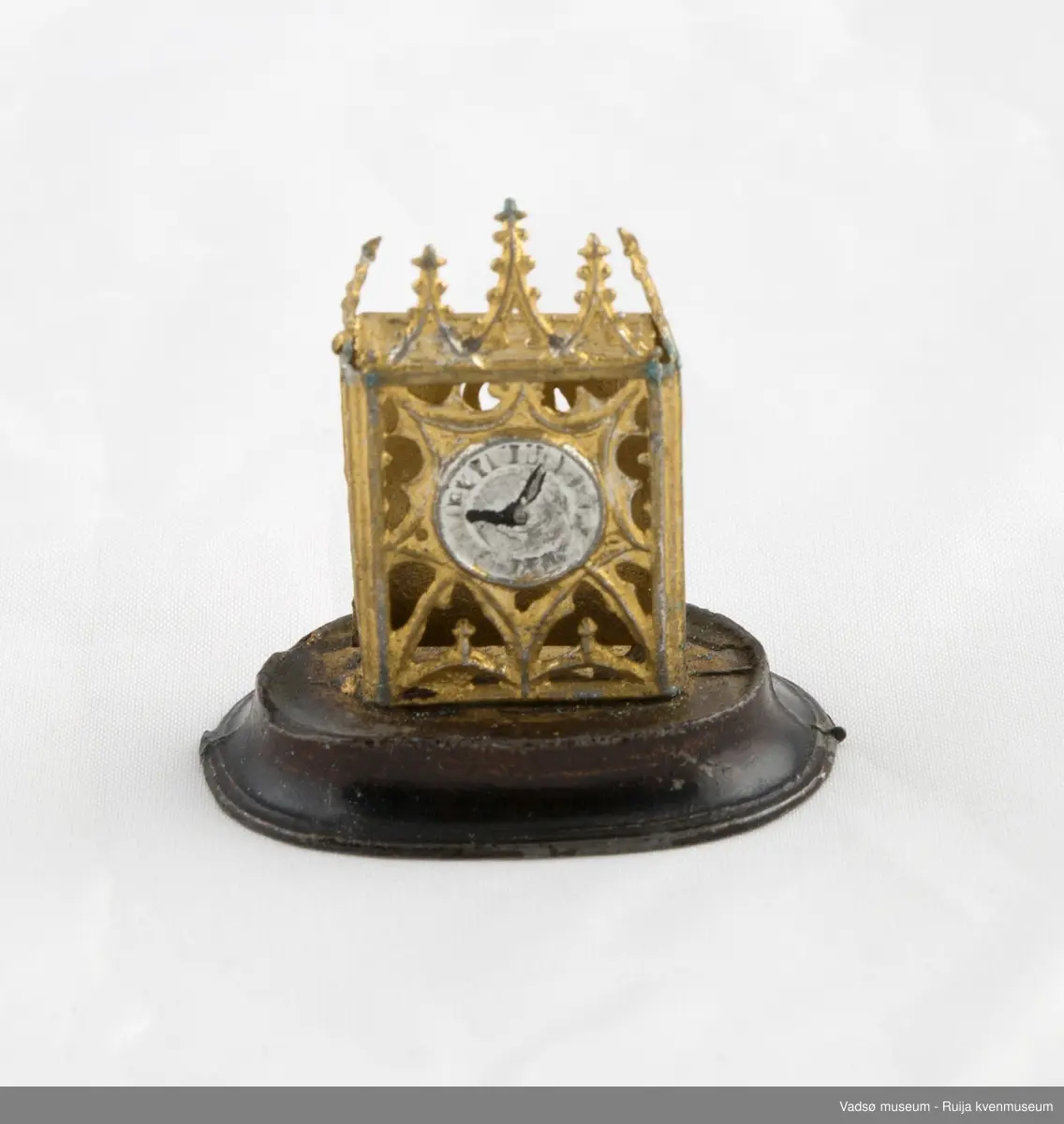 Dukkehusinventar. Forestiller bordklokke i metall. Rektangulært klokkehus med 5 spir i front. Klokkehus er loddet fast i oval metallsokkel som er noe bredere enn klokkehuset. Urskive i sølv og visere i svart. Klokka har en pynte/beskyttelseskuppel i tykt glass som kan tas av og på.
