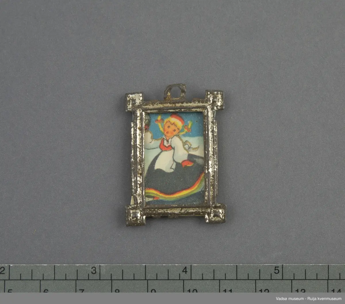 Dukkehusinventar. Bilde med glass og ramme. Rektangulær ramme i sølvfarget tynt metall, utformet i art deco stil. Kvadratisk vulst/ornament i hvert hjørne, øre med rundt opphengshull midt på ene kortsiden. Glasset er festet fast i rammen på baksiden.