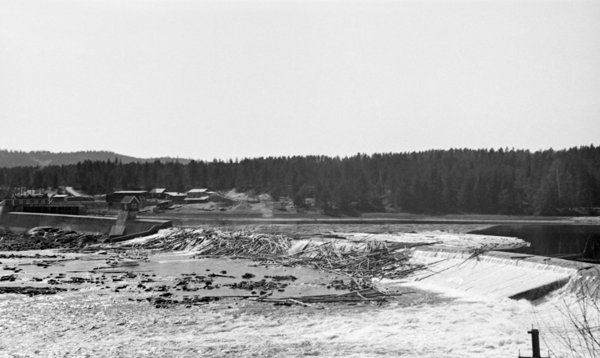 Skjefstadfossen i Glomma, nærmere bestemt i Heradsbygda i Elverum, Hedmark, fotografert i begynnelsen av mai 1944, på et tidspunkt da det var forholdsvis lite vann i elveløpet. Øverst i denne fossen hadde man i 1909-1910 bygd et elvekraftverk med en terskeldam som skulle stuve opp vann til turbinrøret. Overløpsdammen var 312 meter lang, utført i betong og støpt slik at damkrona var noe lavere i den østre enn i den vestre enden. Hensikten var å skape et «sug» i overflata som trakk fløtingstømmeret mot den østre delen av elveløpet. Av samme årsak brukte man å legge ei lense, et flytende stengsel land av sammenkjedete tømmerstokker, som skulle hindre at fløtingsvirket drev inn i inntaksbassenget for vann til kraftstasjonen, som lå i den vestre delen av elveløpet. Da dette fotografiet ble tatt var fløtinga for lengst i gang, men vannføringa hadde avtatt, slik at en god del tømmer hadde blitt liggende i fallsona på den vestre delen av overløpsdammen. Her var det for øvrig bygd en såkalt «tømmerløfter», et skråplan som skulle forebygge at stokkene ble splintret i nedfallssona under dammen.