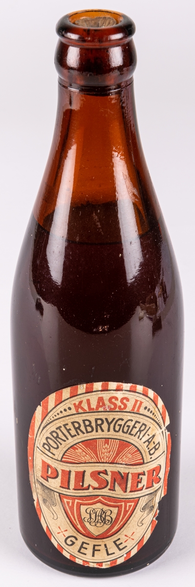 Pilsnerflaska med innehåll. Brunt glas. Etikett: Klass II pilsner, Porterbryggeri AB Gefle.