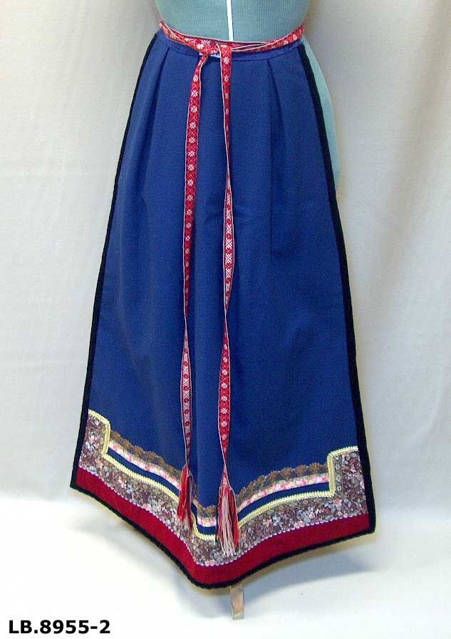 Kan brukast på begge sider, blått på eine sida, lyst på den andre. Beltet skal brettast ned, slik at det framleis viser rettsida ut, når forkleet blir bruka på den blå sida. Forkleet er kanta med blått fløyelsband. Pynta med agramaner i gull og sylv. Band til pynt, ingen broderi. 