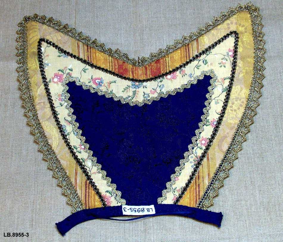  Bringekluten kan brukast på begge sider, raud på den eine, blå og lys gul på den andre. Pynta med agramaner i gull. Band i ymse fargar, ingen broderi. Kan takast av. Hempe nederst framme.
