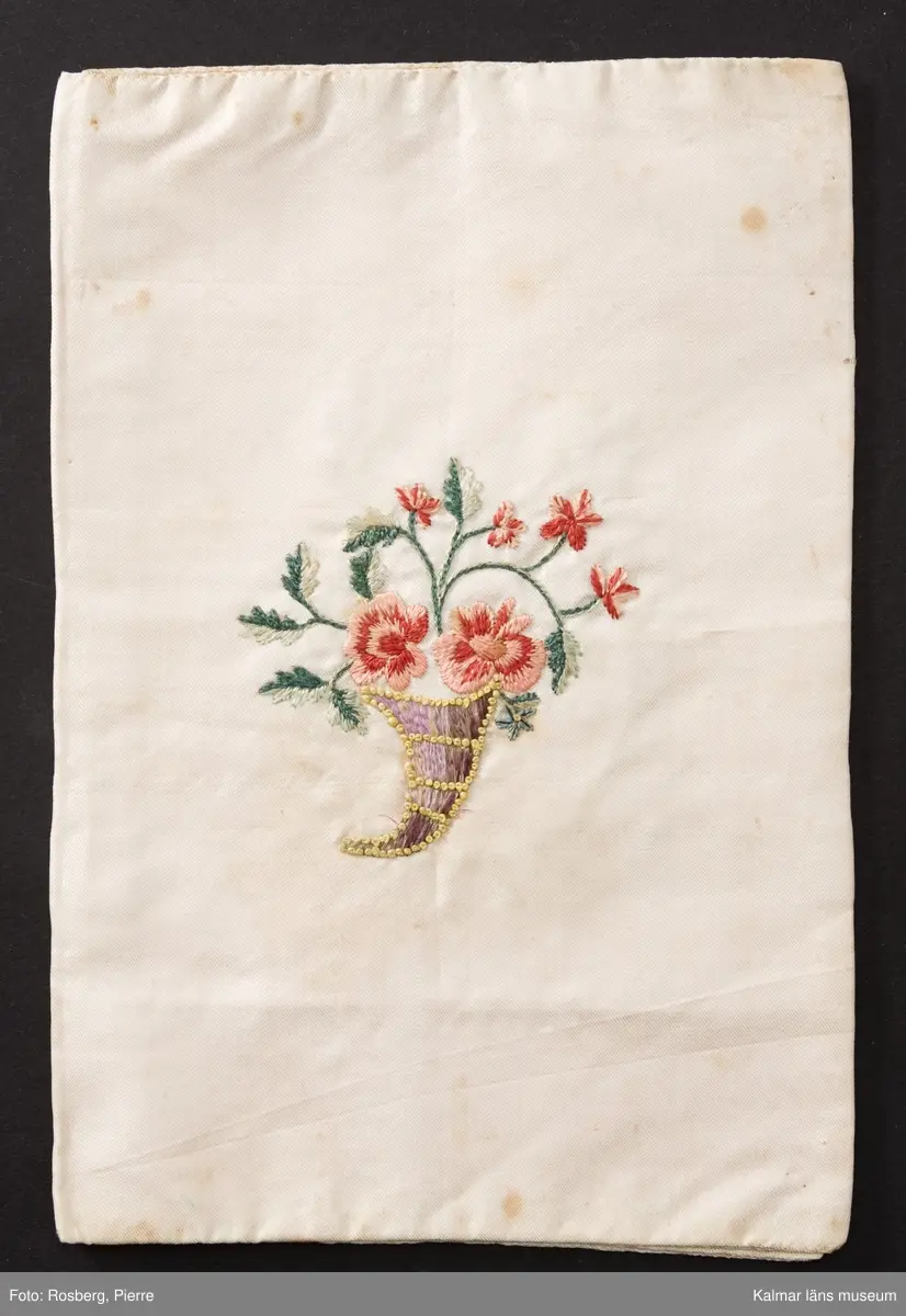KLM 8909. Sedelbok, plånbok. Av vitt siden med broderier av schattérsöm, plattsöm, knutsöm, kjedjesöm. På in- och utsidornas fält blomdekorer. Insidorna märkta inom blomsterkransar med text: M G F 1818. Bröllopsgåva, fästegåva till Magnus Gudmundsson Fröberg.