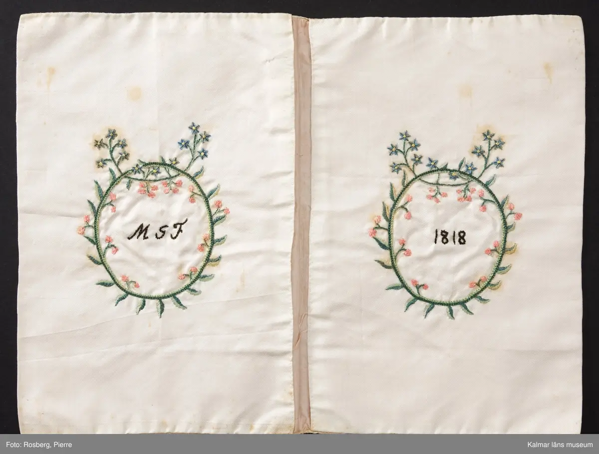 KLM 8909. Sedelbok, plånbok. Av vitt siden med broderier av schattérsöm, plattsöm, knutsöm, kjedjesöm. På in- och utsidornas fält blomdekorer. Insidorna märkta inom blomsterkransar med text: M G F 1818. Bröllopsgåva, fästegåva till Magnus Gudmundsson Fröberg.