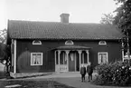 TVÅ SMÅ POJKAR STÅR PÅ GÅRDSPLANEN FRAMFÖR ETT BOSTADSHUS.