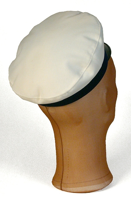 Skärmmössa av vit sammet, lackskärm. Blått sammetsband runt kullen. 
   Mått: Runt kullen 600 mm. Höjd 65 mm. 
   Storlek: 55.