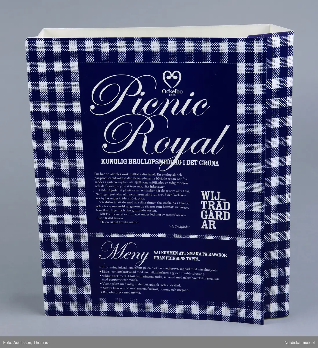 Fodral/förpackning av papp, rektangulär pappskiva vikt för att passa runt besticklåda (NM.0334318), tryckt i blått och vitt efterliknande textil. Text på alla sidor.
Text på framsidan: "Ockelbo 2010 / Picnic Royal / KUNGLIG BRÖLLOPSMIDDAT I DET GRÖNA"..."Allt komponerat och tillagat under ledning av mästerkocken Rune Kalf-Hansen."..."WIJ TRÄDGÅRDAR"..."Meny / Välkommen att smaka på råvaror från prinsens täppa. / Strömming inlagd i granskott på en bädd av svedjerova, toppad med nässelmajonäs. / Rädis- och ärskottsallad emd rökt vildsvinskorv, ägg och tranbärsdressing / Vildsvinsstek med libbstickamarinerad gurka, serverad med nakenhavrekräm smaksatt med pepparrot och vitlök. / Vitmögelost med inlagd rabarber, gräslök- och vildsallad. / Mattes knäckebröd med sparris, färskost, honung och oregano. / Rabarberdryck med mynta.".

På sidorna: "IKEA UTMANAR / Vad kan man mer anvånda en besticklåda till? Lämna ditt förslag vid tennisbanan på Wij Trädgårdar coh var med om utlottningen av en festdukning. Se vinster och lämna svar senast den 19 juni.!"

På baksidan text om de olika matproducenterna:
"Vi är storlta över våra producenter och leverantörer"..."Vildsvin från Hagsta Vilt"..."Honung från Ockelbo"..."Ägg från Axlunds ägg, Mörtebo"..."Nakenhavre från Warbro Kvarn, Katrineholm"..."Strömming från Bergmans Fisk, Norrsundet"..."Sparris och rabarber från gården Böras i Gavelhyttan"..."Kallpressad rapsolja och ärkskott från gården Nygårds i Ovansjö"..."Lagrad vitmögelost från Eskesta Gårdsmejeri, Tierp"..."Rädisor, majrova coh svedjerova från Västerbo, Ockelbo"..."Granskott och tranbär från Lottaboden, Los"..."Vild sallat från Wij Tråädgårdar, Ockelbo"..."Crème fraiche coh youghurt från Gefleortens mejeri"..."'Knäckebröd från Mattes Bröd, Ockelbo"...
/Leif Wallin 2015-01-08