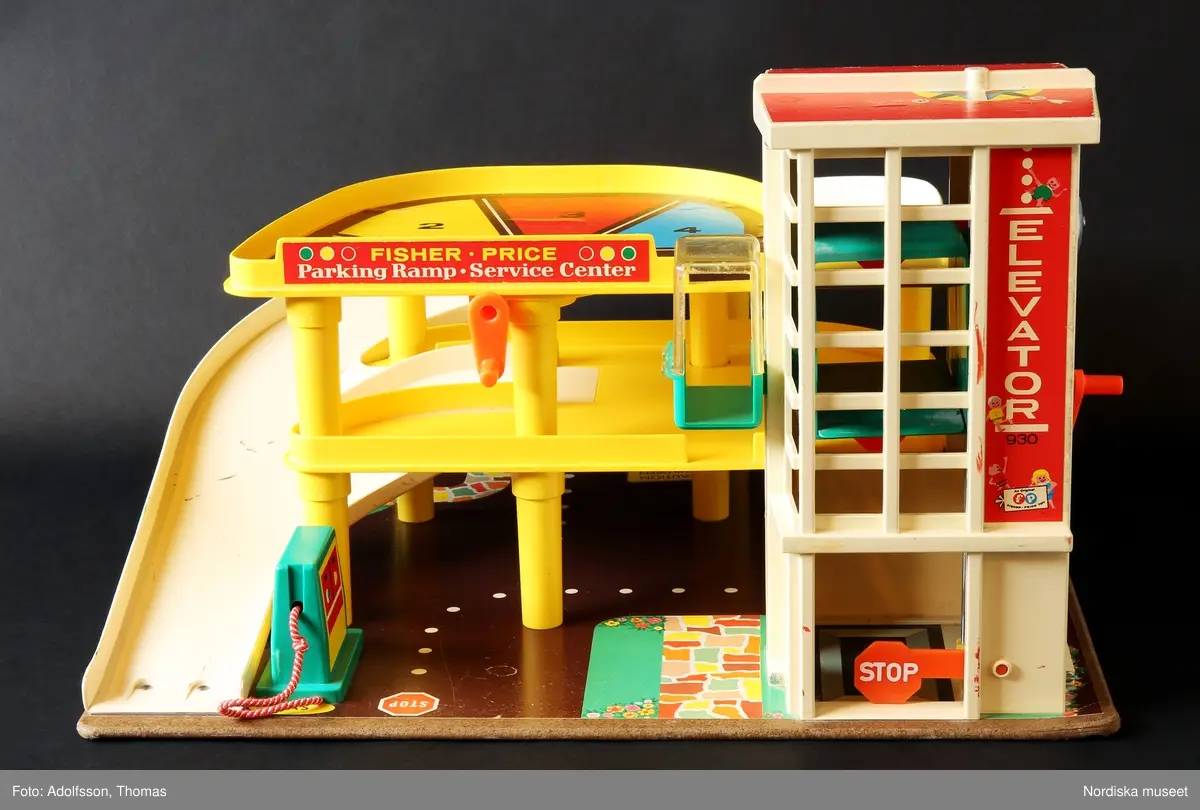 a) Garage med ramp och parkeringsplatser i två våningar för leksaksbilar. Formgjuten plast i klara färger, med påklistrade pappersetiketter med text och bild. Enkel mekanik för att höja eller sänka en hiss för tillhörande leksaksbilar med människofigurer samt roterande platta på det övre planet. Garaget står på en platta av masonit. På nedre plan finns en fast bensinpump med slang av textilt material samt b) en lös ramp med mekanik för höjning och sänkning av leksaksbilar. Till garaget hör fyra bilar som kan rulla nedför en ramp från parkeringsplatserna på det översta planet.