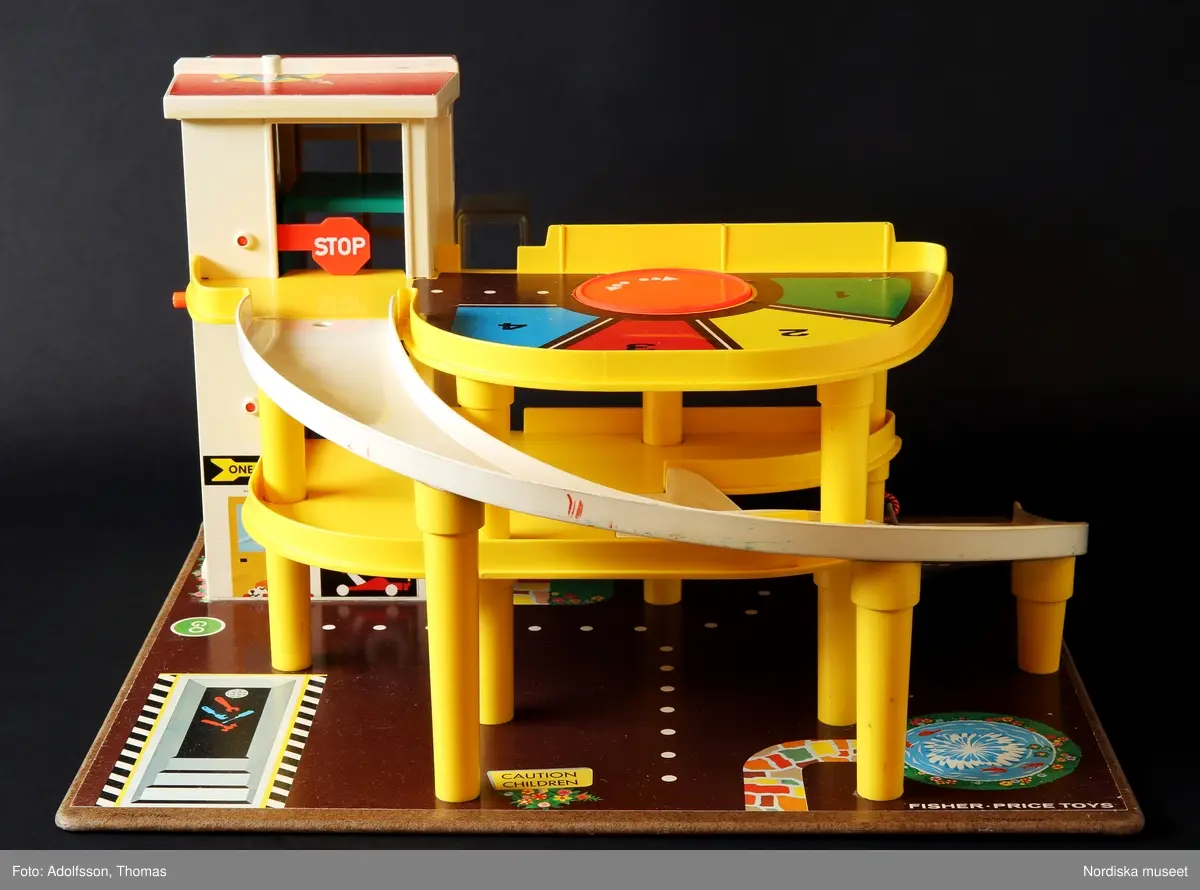 a) Garage med ramp och parkeringsplatser i två våningar för leksaksbilar. Formgjuten plast i klara färger, med påklistrade pappersetiketter med text och bild. Enkel mekanik för att höja eller sänka en hiss för tillhörande leksaksbilar med människofigurer samt roterande platta på det övre planet. Garaget står på en platta av masonit. På nedre plan finns en fast bensinpump med slang av textilt material samt b) en lös ramp med mekanik för höjning och sänkning av leksaksbilar. Till garaget hör fyra bilar som kan rulla nedför en ramp från parkeringsplatserna på det översta planet.