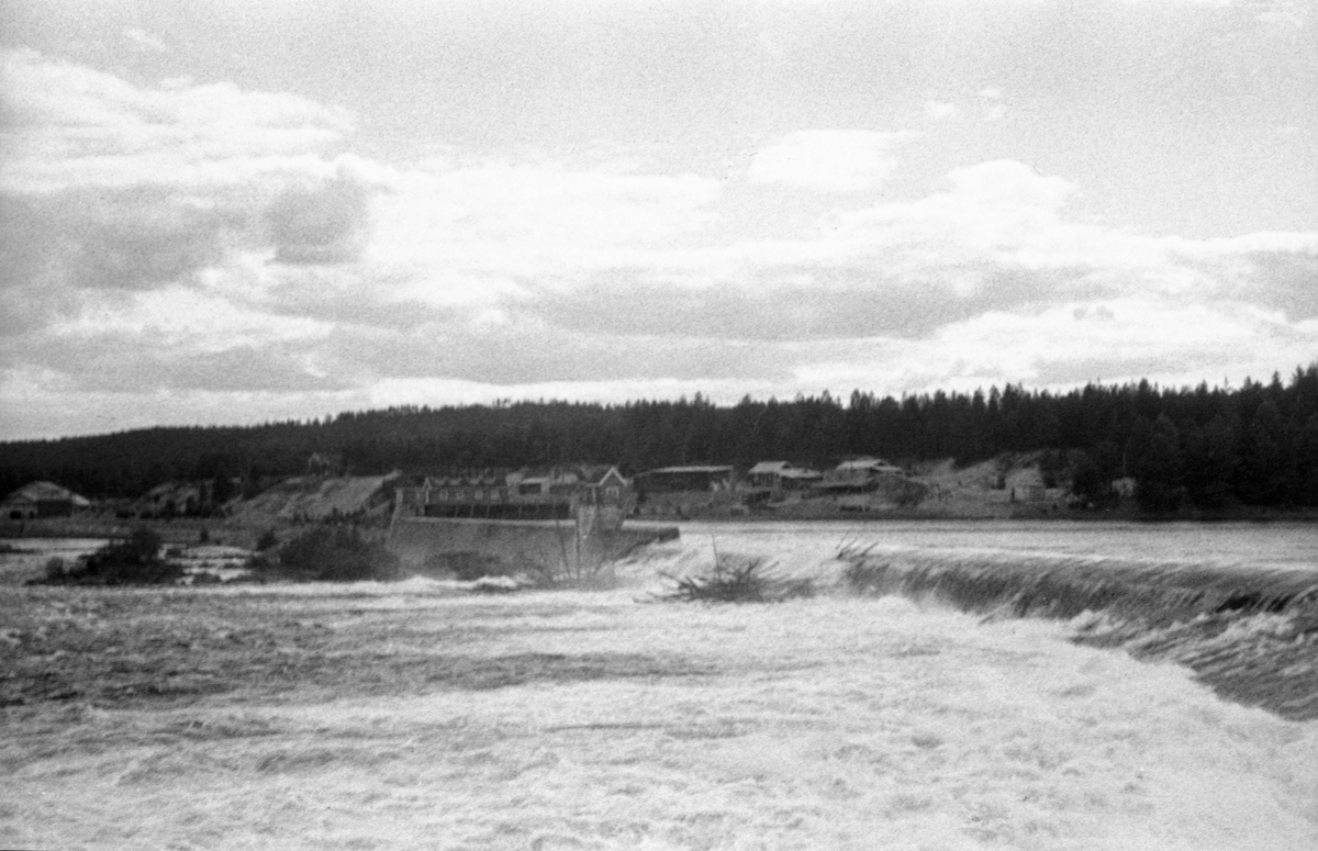 Skjefstadfossen i Glomma i Heradsbygda i Elverum, fotografert i midten av juni i 1946. På dette stedet ble det første store kraftverket i den øvre delen av Glomma bygd i 1909-1910.  I samband med dette prosjektet ble det støpt en 312 meter lang terskeldam på tvers av elveløpet. Denne konstruksjonen skulle stuve opp vann i den ovenforliggende delen av elva og sørge for kontinuerlig tilsig til kraftverket inntaksdam ved den vestre elvebredden, til venstre i dette bildet. Terskeldammen ble gjort noe lavere mot den østre enden. Poenget med dette var å skape et strømdrag som trakk mesteparten av fløtingstømmeret mot den østre delen av Skjefstadfossen, der elva var djupere enn i midtsona, hvor det hadde lett for å bygge seg opp digre hauger med fløtingstømmer.  Da dette fotografiet ble tatt lå det en del tømmer under og på den vestre delen av overløpsdammen. Bakenfor, forholdsvis sentralt i bildet, ser vi inntaksdammen for turbinvannet til kraftstasjonen, med lukehus på damkrona.