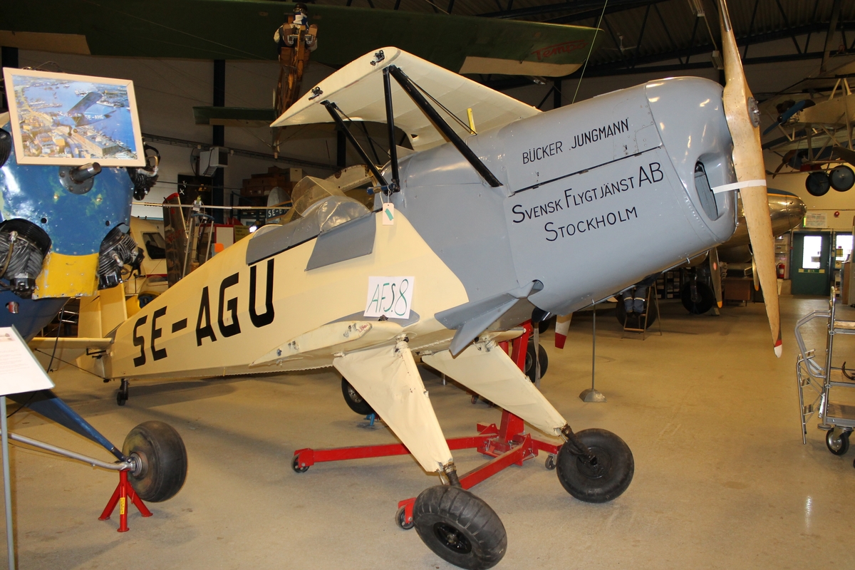 Flygplan av modellen Bücker 131 Jungmann. Dubbeldäckat propellerflygplan, ljusgul- och gråmålat. Två öppna sittbrunnar i tandem och fast landställ.