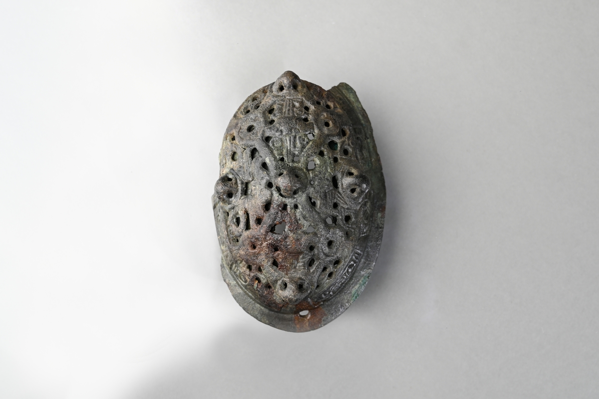 Spännbuckla (se Petersen fig 51) med genombruten överskålla med fem knoppar. Överskållan hel, separerad från underskållan. Underskållan skadad, ornerad längs kanten. Nålfäste och nålhållare kvar, men ej nålen. Tygavtryckmärken från gjurtformen på undersidan, troligen från linne vävt i tuskaft.