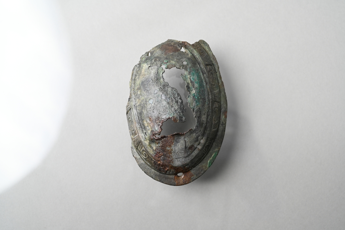 Spännbuckla (se Petersen fig 51) med genombruten överskålla med fem knoppar. Överskållan hel, separerad från underskållan. Underskållan skadad, ornerad längs kanten. Nålfäste och nålhållare kvar, men ej nålen. Tygavtryckmärken från gjurtformen på undersidan, troligen från linne vävt i tuskaft.