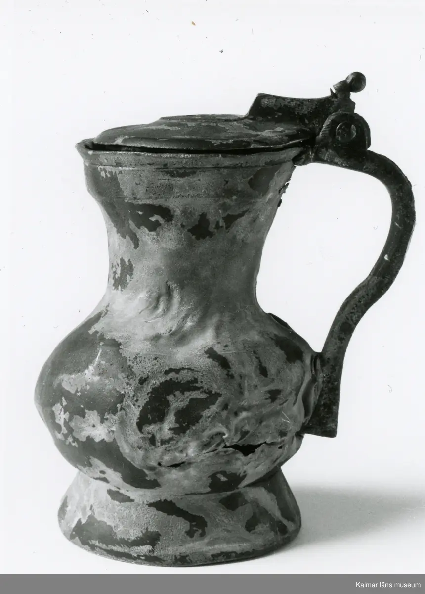 KLM 46428 Kanna av tenn. Bukig form. Hankkärl, försedd med lock och handtag, vidgad botten neråt. I botten, invändigt en medalj med korsfästelsebild. Medaljens kant försedd med pärlring. Ägarmärke/bomärke på lockets och bottens utsidor. Handtaget ornerat med drakmotiv och bladslinga, tumgreppet saknar ett av ekollonen. Kannan skadades svårt vid upphittandet, genomgående sprickor på många ställen.