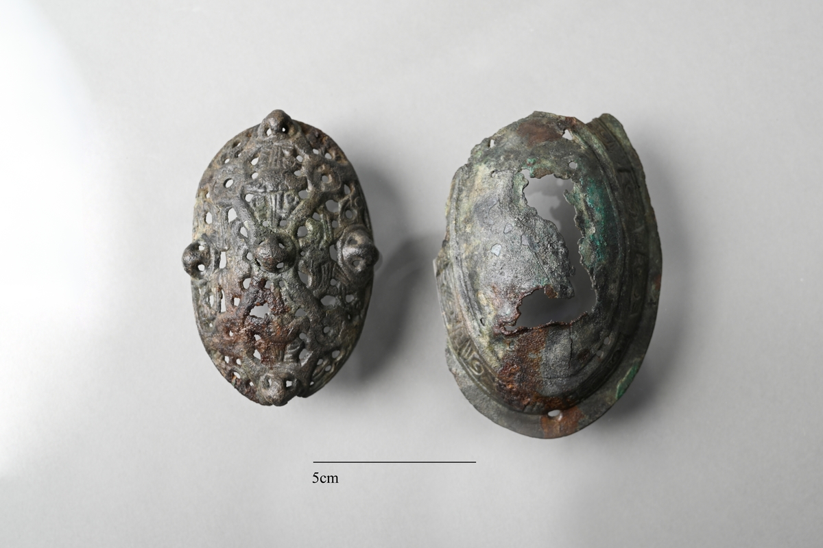 Spännbuckla (se Petersen fig 51) med genombruten överskålla med fem knoppar. Överskållan hel, separerad från underskållan. Underskållan skadad, ornerad längs kanten. Nålfäste och nålhållare kvar, men ej nålen. Tygavtryckmärken från gjurtformen på undersidan, troligen från linne vävt i tuskaft.