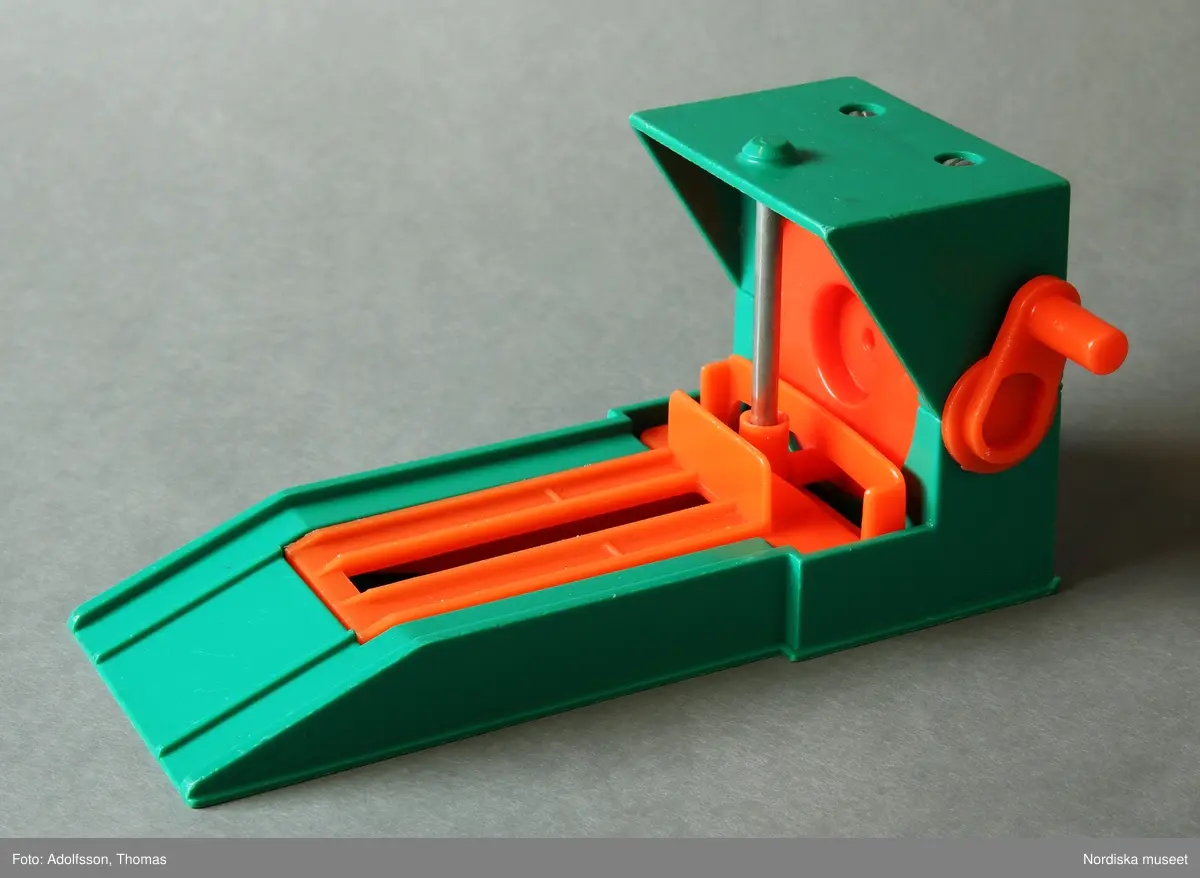 a) Garage med ramp och parkeringsplatser i två våningar för leksaksbilar. Formgjuten plast i klara färger, med påklistrade pappersetiketter med text och bild. Enkel mekanik för att höja eller sänka en hiss för tillhörande leksaksbilar med människofigurer samt roterande platta på det övre planet. Garaget står på en platta av masonit. På nedre plan finns en fast bensinpump med slang av textilt material samt b) en lös ramp med mekanik för höjning och sänkning av leksaksbilar. Till garaget hör fyra bilar som kan rulla nedför en ramp från parkeringsplatserna på det översta planet.