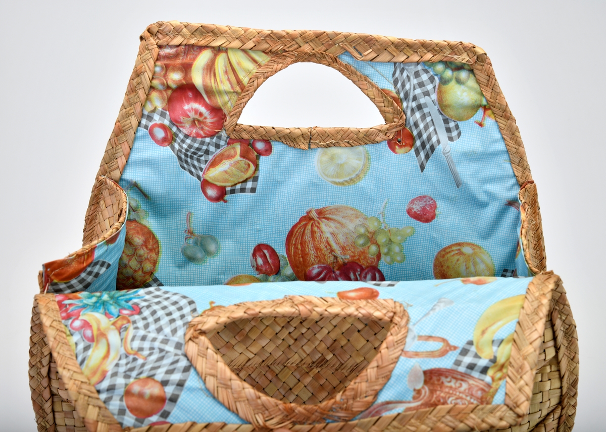 En veske av bast og plast som er flettet og maskinsydd. Basten er flettet. I front (på begge sider) er det 10 vannrette striper som er omtrent 4cm brede. Stripene blir kortere fra øverst og ned mot bunnen (den nederste stripen er kun en liten trekant). På hver side er det 3 skråstilte striper som er flettet med mer "volum". Det er trekantformede håndtak øverst på begge sider. Sidene og bunnen på vesken består av 4 flettede striper. Vesken er foret med plast. Den er blå- og hvitrutet med bilder av diverse frukt og en primus på rutete duk. Mønsteret er repeterende.