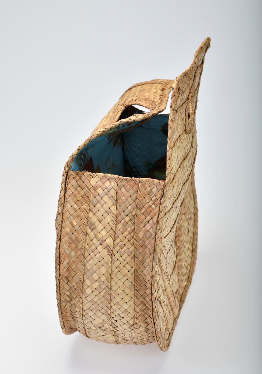 En veske av bast og plast som er flettet og maskinsydd. Basten er flettet. I front (på begge sider) er det 10 vannrette striper som er omtrent 4cm brede. Stripene blir kortere fra øverst og ned mot bunnen (den nederste stripen er kun en liten trekant). På hver side er det 3 skråstilte striper som er flettet med mer "volum". Det er trekantformede håndtak øverst på begge sider. Sidene og bunnen på vesken består av 4 flettede striper. Vesken er foret med plast. Den er blå- og hvitrutet med bilder av diverse frukt og en primus på rutete duk. Mønsteret er repeterende.