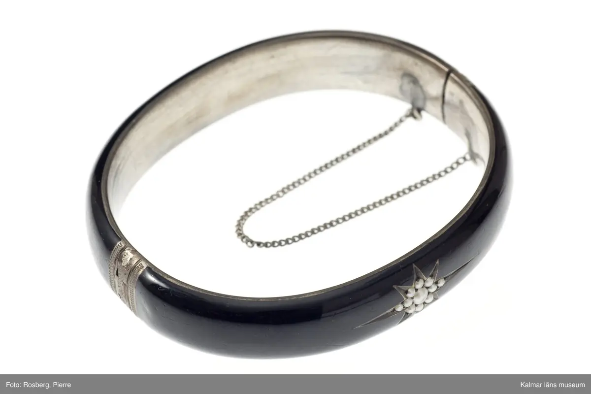 KLM 34780:1  Armring, av svart material. Oval, inläggningar av silver, infattningar av gul metall och pärlor. Förvaras i oval röd ask med text: A Michelsen, Hof og Ordens- Juveler, Kongens Nytorv. Har troligen tillhört Jenny Nyström.