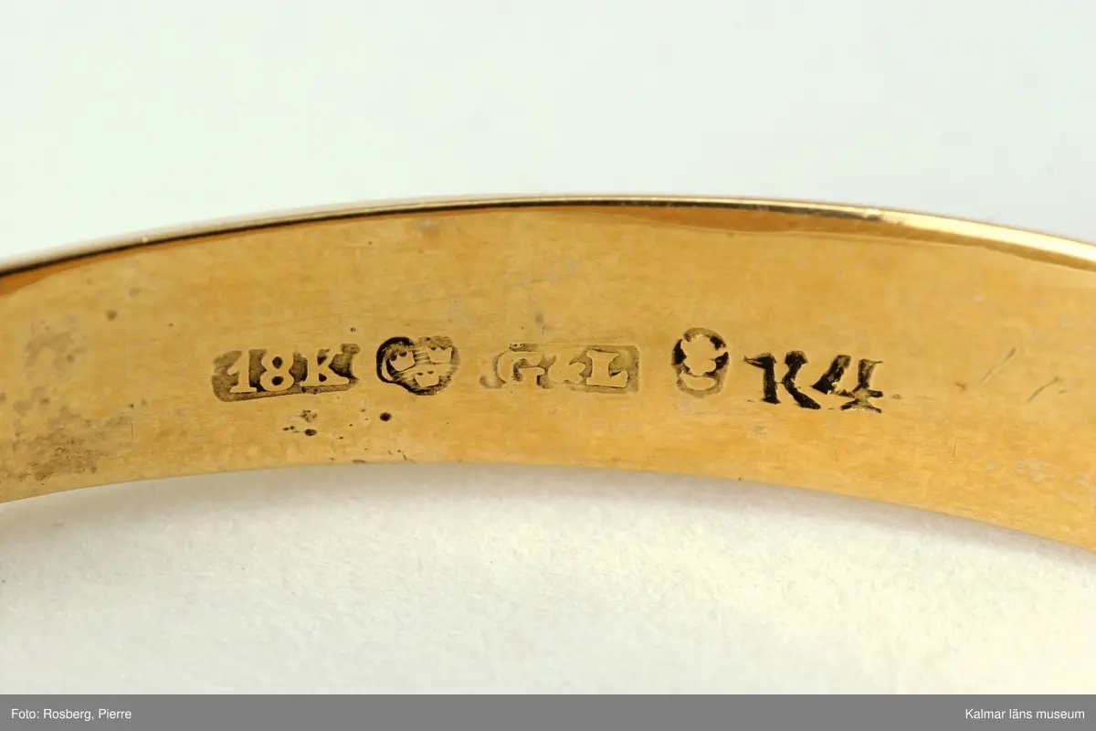 KLM 34781 Armring av guld. Oval form. Ihålig armring, slät undersida, ovansidan rikt dekorerad med blad och blommönster. Förvaras i fyrkantig pappask, gulfärgad, tillsammans med gul hårlock. Stämplar: 18K, kontrollstämpel, mästarstämpel, Giron & Löngren (1820-1847), stadsstämpel, Stockholm, årsstämpel, K4 =1840. Inskription: d 15 juni och d 9 juli 1840.