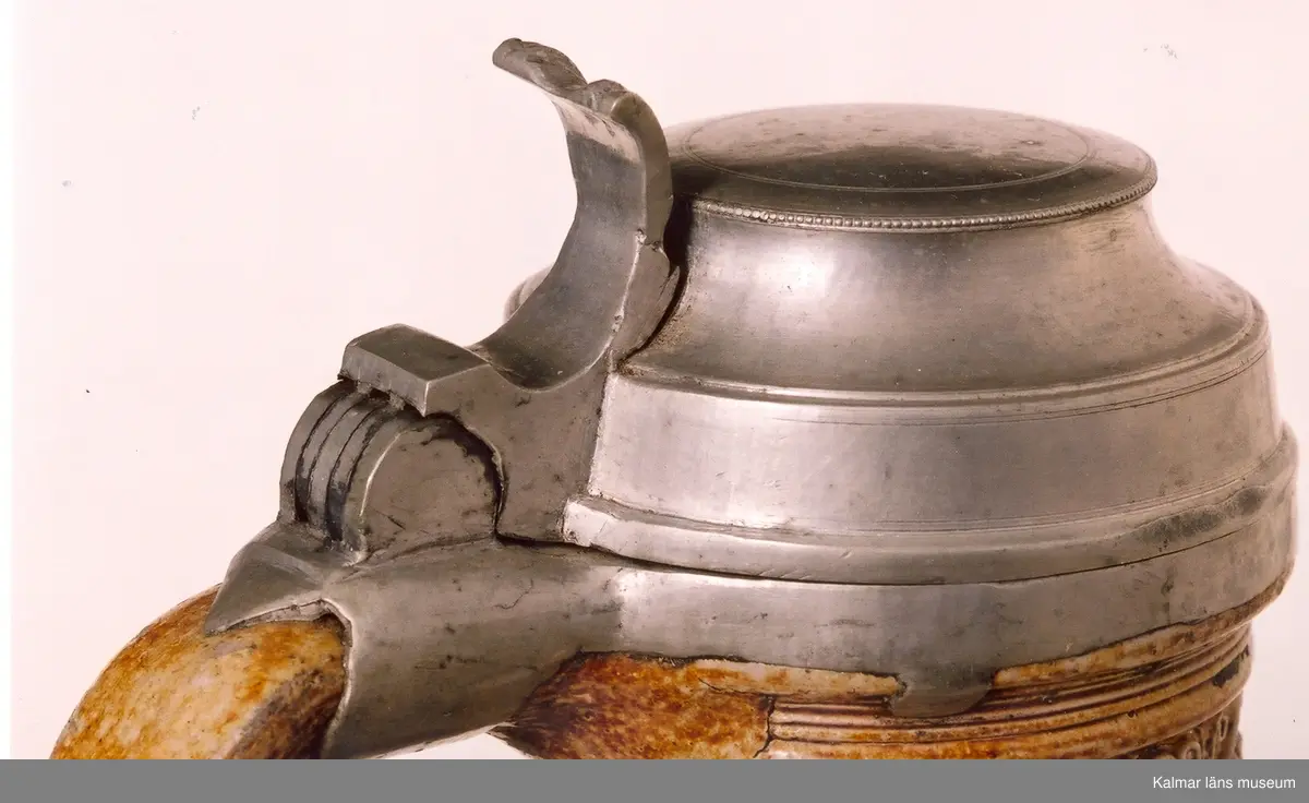 KLM 1712. Krus av stengods. Saltglaserat. Bukigt. Krus med hänkel. Lock och fot av tenn. Tumgrepp: klogrepp. Rhenkrus med bandinskription och årtalet 1583. Inskription: Geret du mus daper blasen, so dansen die buren, als waren se rasen, fri uf, spricht pastor, ich ver dans, di kap mit den kor. Reliefmodelerad dekor. Keramik av Siegburgtyp, Tyskland.