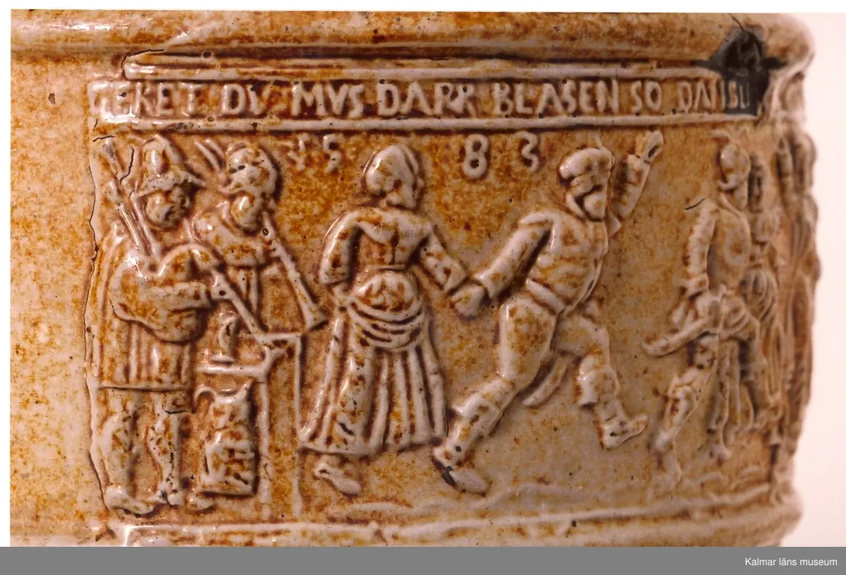 KLM 1712. Krus av stengods. Saltglaserat. Bukigt. Krus med hänkel. Lock och fot av tenn. Tumgrepp: klogrepp. Rhenkrus med bandinskription och årtalet 1583. Inskription: Geret du mus daper blasen, so dansen die buren, als waren se rasen, fri uf, spricht pastor, ich ver dans, di kap mit den kor. Reliefmodelerad dekor. Keramik av Siegburgtyp, Tyskland.