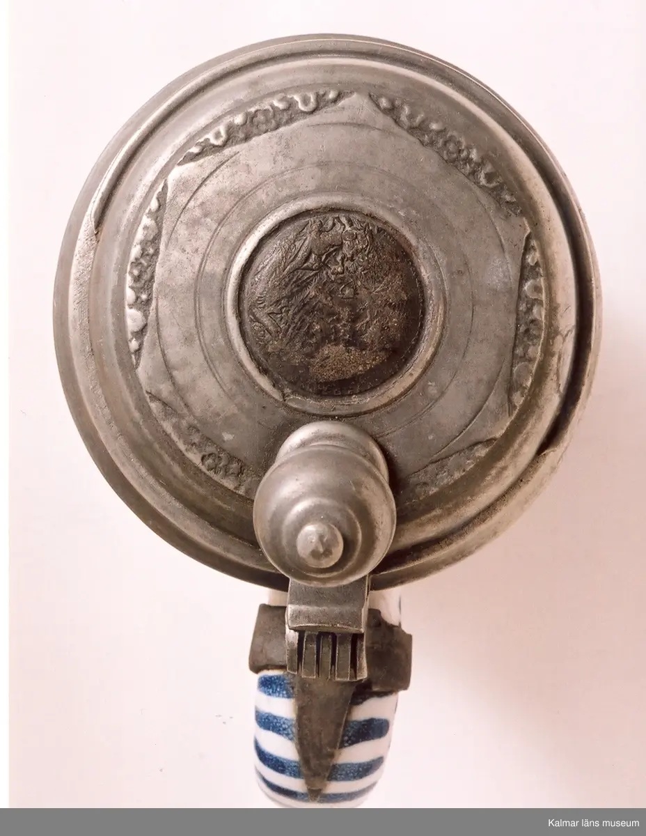 KLM 5725. Krus av fajans. Cylindrisk med grepe och tennlock, vari avbildning av en tysk medalj. På kruset ett landskap med hjort och hund, allt i blått, violett och gult. Tumgrepp: kula med gördel och krönknopp. Tennskodd botten. Tillverkat i Tyskland.