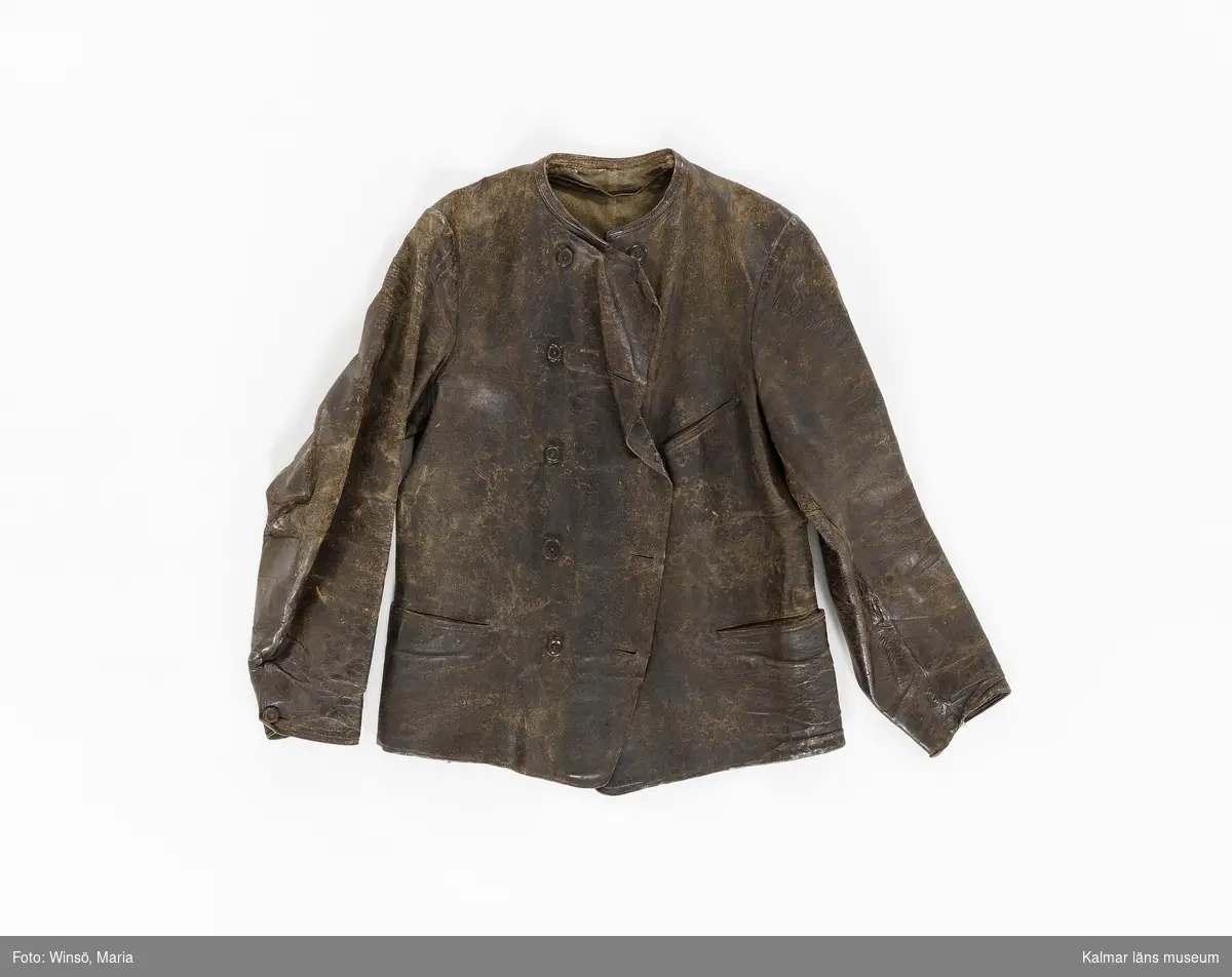 KLM 46056:1. Jacka. Skinnjacka. Arbetsjacka. Använd ytterplagg. Av mörkbrunt skinn. Kort modell. Dubbelknäppning med fem par knappar, alla udda, och knapphål. Utan krage. Dolda fickor fram samt bröstficka på vänster sida. Slejf med spänne i ryggen. Lång ärm som avslutas med kort sprund som knäpps med knapp och knapphål. Grönbrunt foder av kypertvävd bomull/ylle. Fastsydd hank av brun mocka i fodret i nacken.