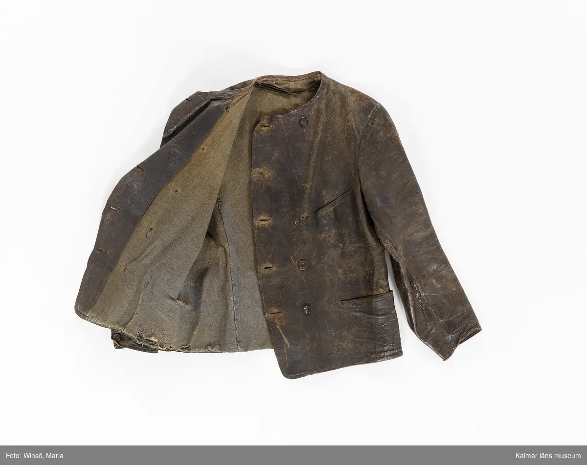 KLM 46056:1. Jacka. Skinnjacka. Arbetsjacka. Använd ytterplagg. Av mörkbrunt skinn. Kort modell. Dubbelknäppning med fem par knappar, alla udda, och knapphål. Utan krage. Dolda fickor fram samt bröstficka på vänster sida. Slejf med spänne i ryggen. Lång ärm som avslutas med kort sprund som knäpps med knapp och knapphål. Grönbrunt foder av kypertvävd bomull/ylle. Fastsydd hank av brun mocka i fodret i nacken.